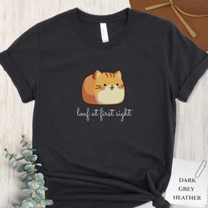 May include: Dark grey heather t-shirt featuring a cartoon cat designed to look like a loaf of bread. The text "loaf at first sight" is written below the cat. The shirt is a casual wear item.