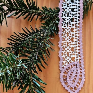 May include: A handmade, white and mauve bobbin lace bookmark with a tassel, displayed with a sprig of evergreen. The bookmark features intricate openwork patterns and a delicate, fringed edge. The image includes the text "bobbin lace patterns by Tina Koder."