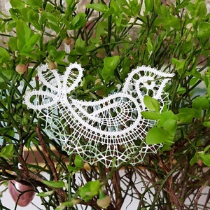 May include: A white bobbin lace bird ornament hangs from a branch of a green bush. The bush is in a clear glass vase. There are several small, pink and green ornaments hanging from the branches.