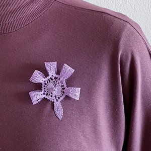 May include: A purple bobbin lace flower brooch with a delicate design. The brooch is pinned to a purple shirt. The text "bobbin lace patterns by TINA KODER" is visible in the background.