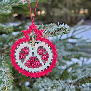 May include: A red and white bobbin lace Christmas ornament suspended from a frosted evergreen branch. The ornament features a circular design with red beads and the text "bobbin lace patterns by TINA KODER".
