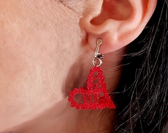 May include: Red heart-shaped bobbin lace earrings with silver hooks. The earrings are worn by a person with dark hair.