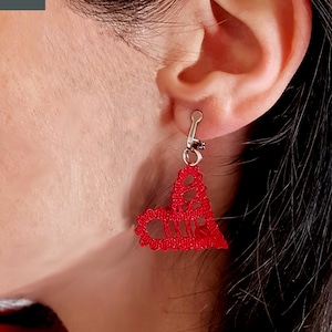 May include: Red heart-shaped bobbin lace earrings with silver hooks. The earrings are worn by a person with dark hair.