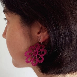 May include: A person wearing a pair of pink bobbin lace earrings. The earrings are in the shape of a flower with a delicate design.