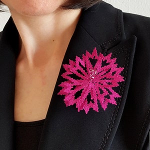 Puede incluir: Un broche de encaje de bolillos de color rosa fijado a una chaqueta negra. El texto "bobbin lace patterns by TINA KODER" es visible en la esquina inferior derecha de la imagen.