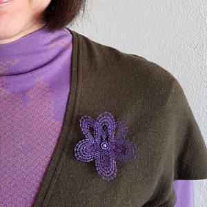 May include: A purple bobbin lace flower brooch with a beaded center, pinned to a purple shirt and brown cardigan. The image includes the text "bobbin lace patterns by TINA KODER".