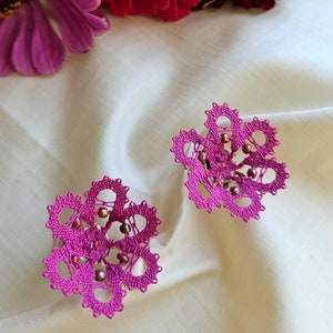 May include: Two delicate pink bobbin lace flowers with a beaded center. The lace is handmade and intricate. The flowers are on a white background.