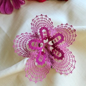 May include: A pink bobbin lace flower with a delicate design and beaded accents. The lace is displayed on a white fabric background with pink and red flowers.