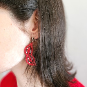 Pode incluir: Apresenta brincos de renda de bilros vermelhos com detalhes em preto e branco. Os brincos, em forma de folha, estão presos a um gancho dourado. A imagem também inclui o texto "bobbin lace patterns by TINA KODER."