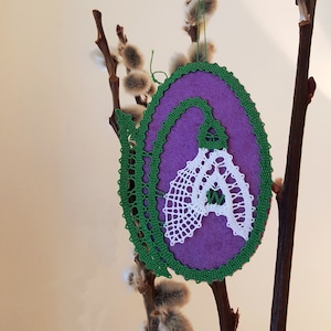 Può includere: Un ornamento in pizzo a fuselli di colore viola e verde a forma di bucaneve. L'ornamento è appeso a un ramo con boccioli bianchi e pelosi.