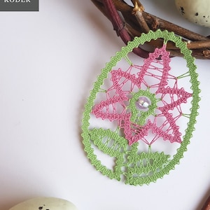 May include: A handmade bobbin lace Easter egg ornament. The egg-shaped ornament features a pink flower design within a green border, accented with a clear gem. The background includes speckled eggs, twigs, and the text "bobbin lace patterns by TINA KODER."