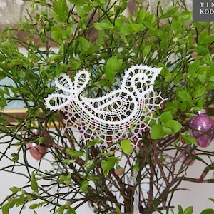 May include: A white bobbin lace bird design with a light pink and green gradient, displayed in a vase with green leaves and pink and green glass ornaments. The text "bobbin lace patterns by TINA KODER" is visible in the upper right corner of the image.