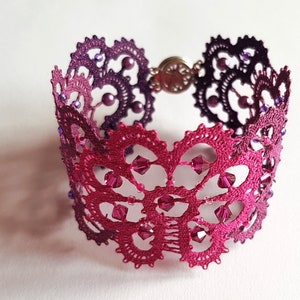 May include: A purple and pink bobbin lace bracelet with a clasp closure. The bracelet is decorated with small beads and crystals. "bobbin lace patterns by TINA KODER" is printed on a gray background.