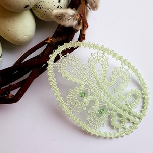 May include: A light green bobbin lace oval with a floral design. The lace is displayed on a nest of twigs and speckled eggs. The text "bobbin lace patterns by TINA KODER" is in the upper right corner of the image.