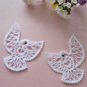 May include: Two white bobbin lace dove ornaments with black bead eyes are displayed on a pink surface. The doves are near a bouquet of white, yellow, and purple flowers. The image also includes a logo that says "bobbin lace patterns by TINA KODER."