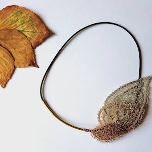 May include: A delicate gold and copper leaf-shaped bobbin lace necklace with a black cord and a gold metal bar. The necklace is displayed on a white surface with a dried leaf.
