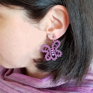 May include: Purple bobbin lace earrings with a butterfly design and small purple beads. The earrings are worn by a person with dark hair.