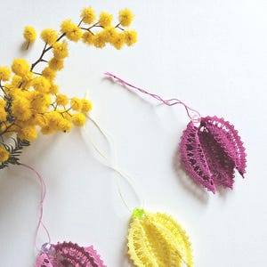 May include: Three handmade bobbin lace flower charms in pink, yellow, and a gradient of pink. The charms are hanging from white thread and have a small bead at the top. The background is white.