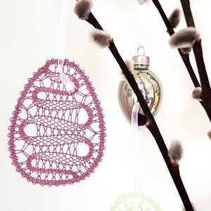May include: Two delicate, handmade lace Easter eggs, one pink and one green, hang from willow branches with pink and green glass ornaments. The text "Tina Koder original idrija lace design" is visible in the bottom left corner.