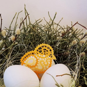 Puede incluir: Dos huevos blancos y un pájaro de ganchillo amarillo en un nido de ramitas verdes y brotes blancos y esponjosos. El texto "TINA KODER original idrija lace design" está en la esquina inferior derecha.