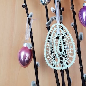 May include: Four Easter eggs hanging from willow branches. One egg is a light green lace design, one is a silver glass ornament, and two are purple glass ornaments.