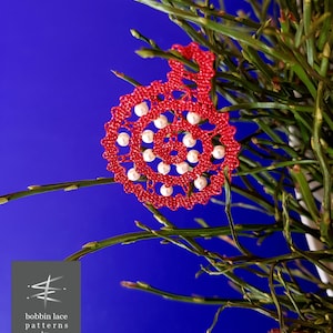 May include: A red bobbin lace ornament with white pearl beads, against a blue background. The ornament is in the shape of a spiral. The text "bobbin lace patterns by TINA KODER" is visible in the lower left corner of the image.