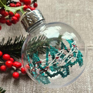 May include: A clear glass Christmas ornament with a white and teal lace design inside. The ornament has a silver top and is decorated with red and white beads.