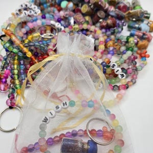 Peut inclure: Une collection de bracelets et porte-clés perlés colorés. Un sac en filet blanc contient un bracelet avec l'inscription "WISH" et une petite bouteille. Les bracelets et porte-clés sont de différentes couleurs, dont le rose, le bleu, le vert et le jaune.