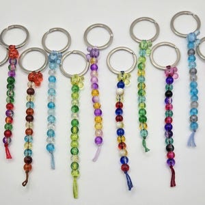 Könnte beinhalten: Eine Sammlung von Schlüsselanhängern mit Perlen in verschiedenen Farben. Jeder Schlüsselanhänger hat einen silbernen Ring und eine Kette aus bunten Perlen, darunter Gelb, Rot, Grün, Blau und Lila. Einige Schlüsselanhänger haben Quasten am unteren Ende.