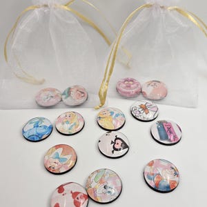 May include: A collection of round, glass-covered magnets featuring characters from Alice in Wonderland. The magnets are in a variety of colors and designs, and are displayed in small, sheer white bags with gold ribbon ties.