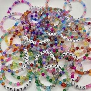 Könnte beinhalten: Eine Sammlung bunter Perlenarmbänder. Jedes Armband hat eine Vielzahl von Perlen in Rosa-, Lila-, Grün-, Orange- und Blautönen. Einige Armbänder haben weiße Buchstabenperlen mit Wörtern wie "WISH", "DREAM" und "MAGIC".