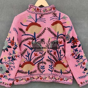May include: A pink embroidered jacket with floral patterns in shades of blue, red, and green. The jacket has a high neck and long sleeves. The text "Crafts Aura Handmade With Love and Care" is embroidered on the back.