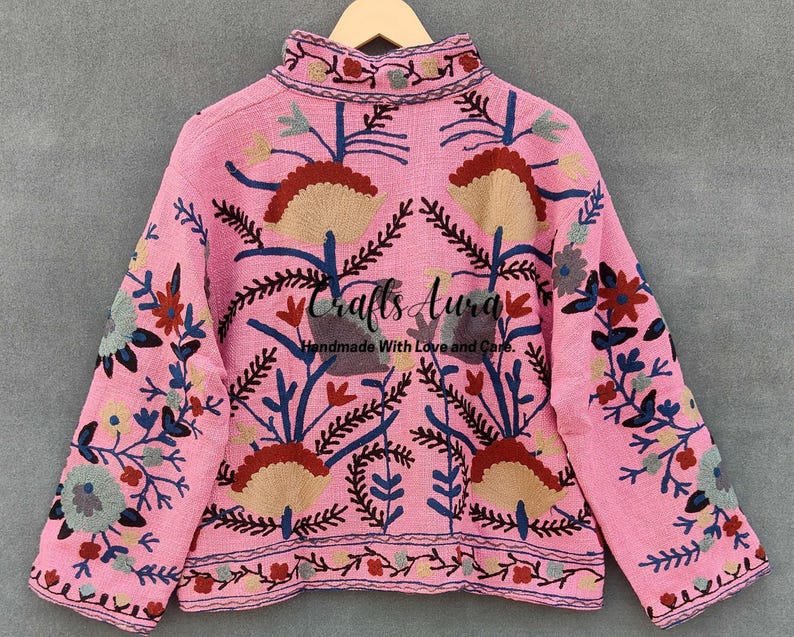 May include: A pink embroidered jacket with floral patterns in shades of blue, red, and green. The jacket has a high neck and long sleeves. The text "Crafts Aura Handmade With Love and Care" is embroidered on the back.