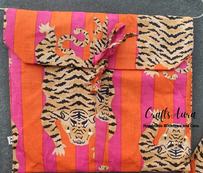 May include: Orange and pink striped fabric with a tiger print. The fabric is folded into a pouch with a pink ribbon tie. The text "Crafts Aura Handmade With Love and Care." is printed on the fabric.