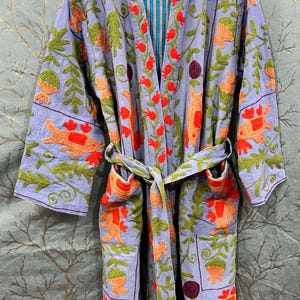 Op de afbeelding: Een lichtpaarse geborduurde badjas met oranje en groene bloempatronen. De badjas heeft een bijpassende riem en zakken. De binnenvoering is blauw en wit gestreept. De tekst "Crafts Aura" en "Handmade With Love and Care" zijn zichtbaar.