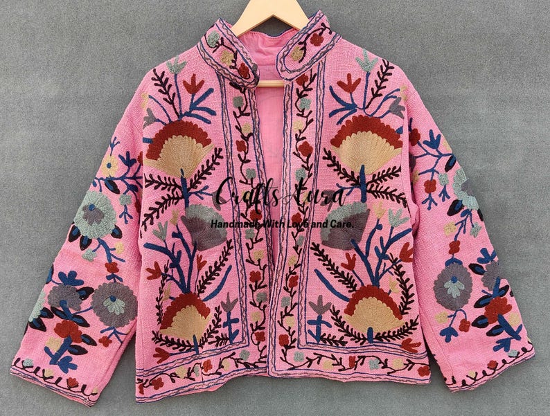 May include: A pink embroidered jacket with floral patterns in shades of blue, red, and beige. The jacket has a collar and a front closure. "Crafts Bazaar Handmade with Love and Care" is written on the jacket.