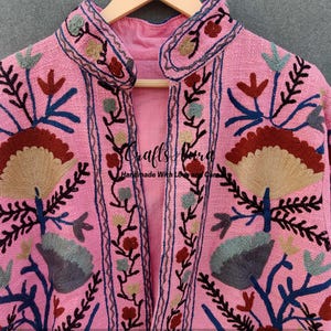 May include: A pink embroidered jacket with a floral design. The jacket has a mandarin collar and a front closure. The embroidery is in shades of brown, blue, and green.