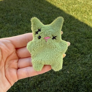 May include: A small, light green plush toy shaped like a creature with pointed ears. It features black embroidered X-shaped eyes, a single black dot for a nose, and a small pink stitched mouth. The texture is fuzzy and soft to the touch.
