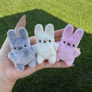May include: Three small plush rabbit toys in gray, white, and light pink. Each rabbit features a fuzzy texture, two upright ears, two small black bead eyes, and a stitched black X-shaped mouth. The figures are compact and soft in appearance.