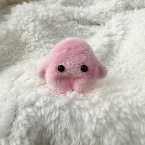 Puede incluir: Un juguete de peluche rosa con ojos negros y una peque&ntilde;a boca. El juguete est&aacute; sentado sobre una superficie blanca y esponjosa.