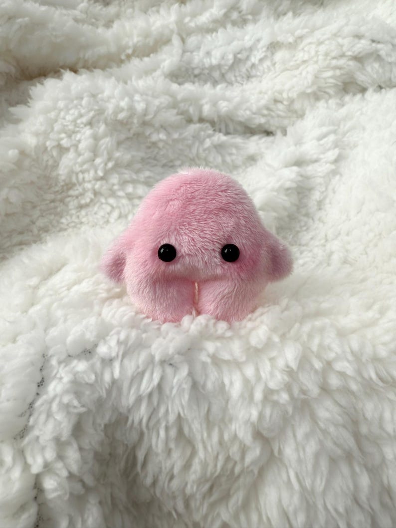 Puede incluir: Un juguete de peluche rosa con ojos negros y una peque&ntilde;a boca. El juguete est&aacute; sentado sobre una superficie blanca y esponjosa.