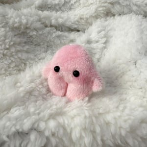 Puede incluir: Un peque&ntilde;o juguete de peluche rosa con ojos negros y cuatro patas rechonchas. El juguete est&aacute; sentado sobre una superficie blanca y esponjosa.