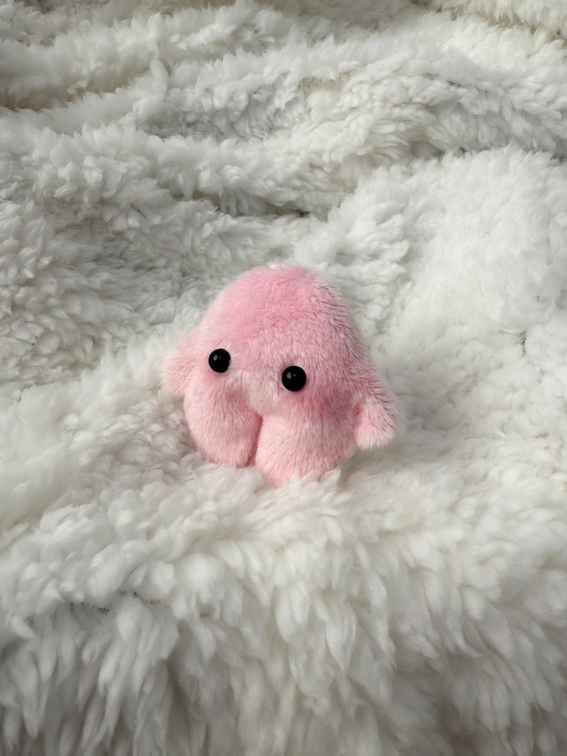 Puede incluir: Un peque&ntilde;o juguete de peluche rosa con ojos negros y cuatro patas rechonchas. El juguete est&aacute; sentado sobre una superficie blanca y esponjosa.
