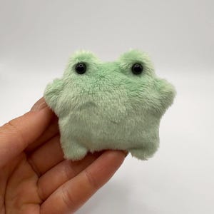 May include: A small, plush green frog with black eyes. The frog is sitting on a hand.