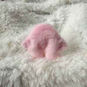 Puede incluir: Un juguete de peluche rosa y esponjoso con forma redondeada y dos peque&ntilde;os brazos. El juguete est&aacute; sentado sobre una superficie esponjosa blanca.