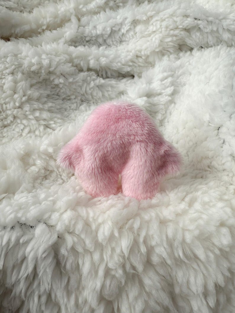 Puede incluir: Un juguete de peluche rosa y esponjoso con forma redondeada y dos peque&ntilde;os brazos. El juguete est&aacute; sentado sobre una superficie esponjosa blanca.