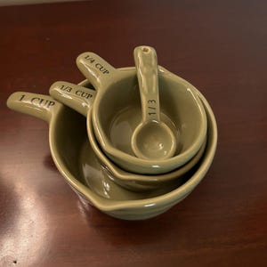 May include: A set of olive green ceramic measuring cups and a measuring spoon nested together. The cups are labeled 1 CUP, 1/3 CUP, and 1/4 CUP, while the small spoon is labeled 1/3. Each piece features a short handle and a smooth, glossy finish.