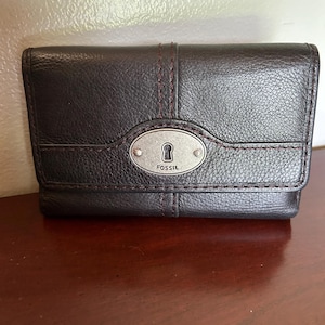 May include: A dark brown textured leather wallet featuring a horizontal flap with a central silver-toned oval metal plaque. The plaque displays a keyhole icon and the word FOSSIL embossed in the center, accented by dark red contrast stitching.