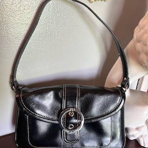 May include: Black shoulder bag with a silver buckle closure. The bag has a rectangular shape with a curved flap and a single shoulder strap. White stitching accents the edges and center of the bag. The brand name "Kathy Van Zeeland" is visible on the buckle.