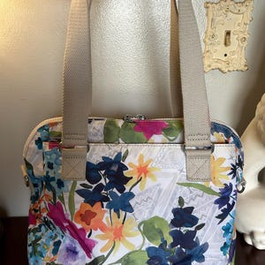 May include: A white handbag with a floral print featuring bright pink, yellow, blue, and green flowers. The bag has two beige shoulder straps.